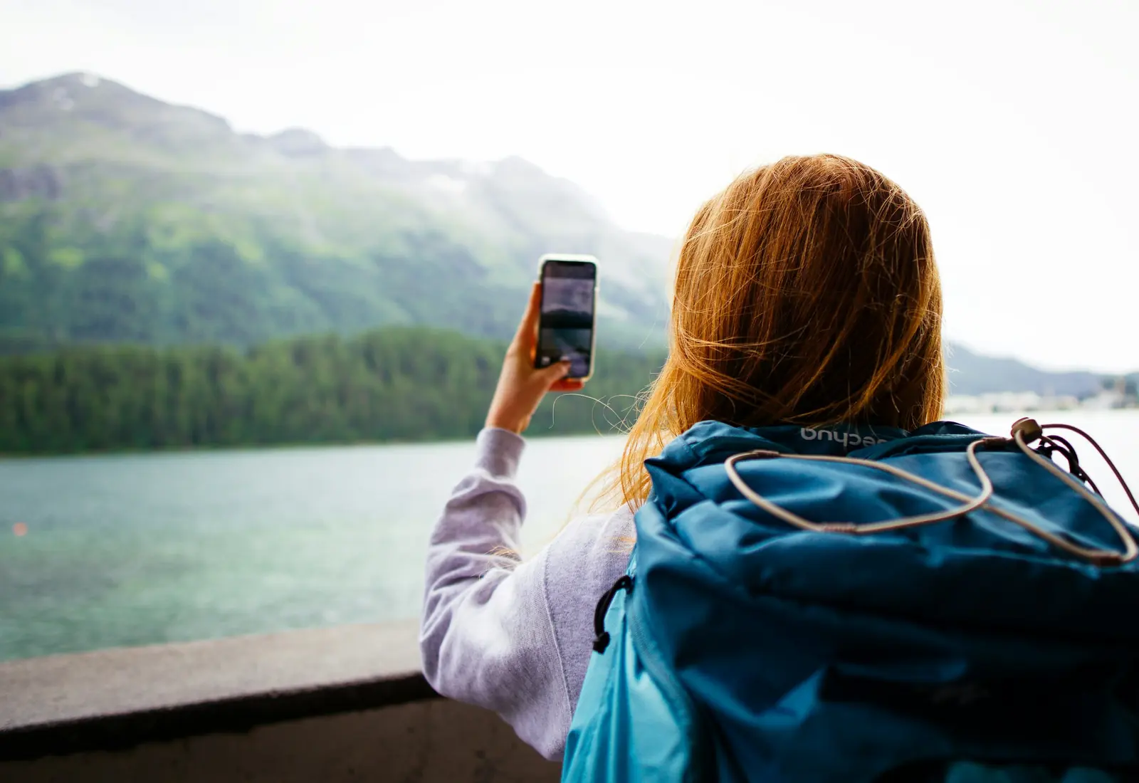 Traveler with a backpack taking a photo of a mountain lake with her phone